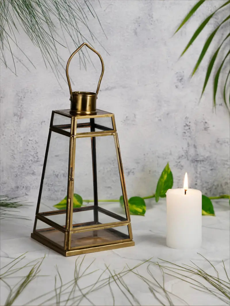 Brass lantern with a white candle on a light gray surface with green leaves in the background