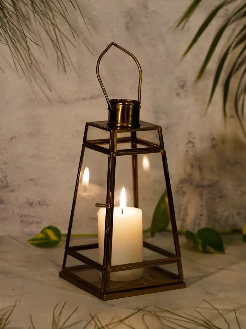 Brass lantern with a lit candle inside, placed on a textured surface with green leaves in the background.