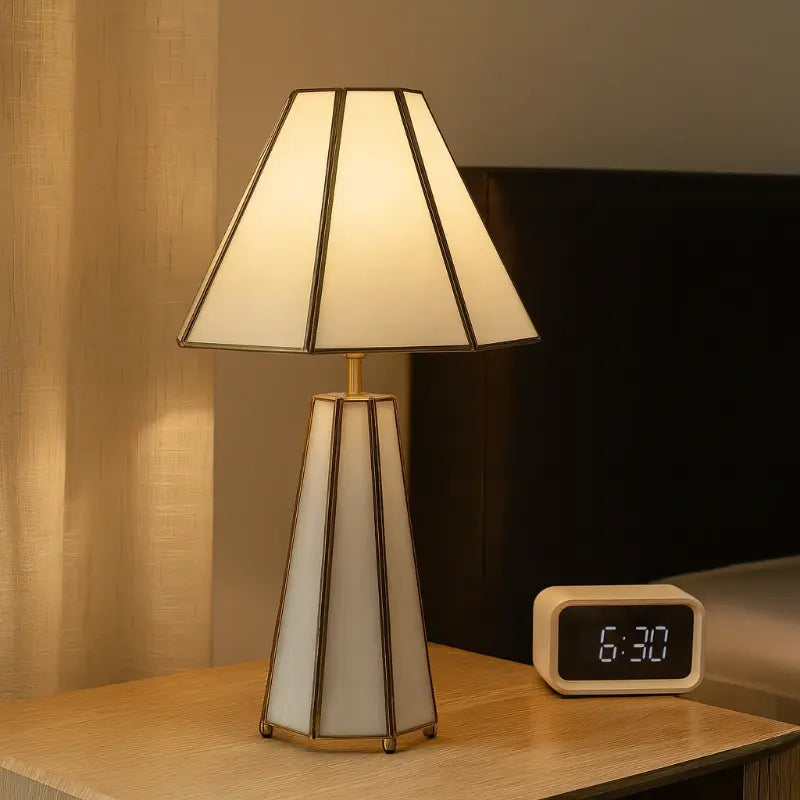 Table lamp with a white shade and gold accents on a wooden surface, with a digital clock displaying 6:30 in the background.