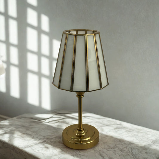 Gold table lamp with a white shade on a marble surface with sunlight casting shadows.